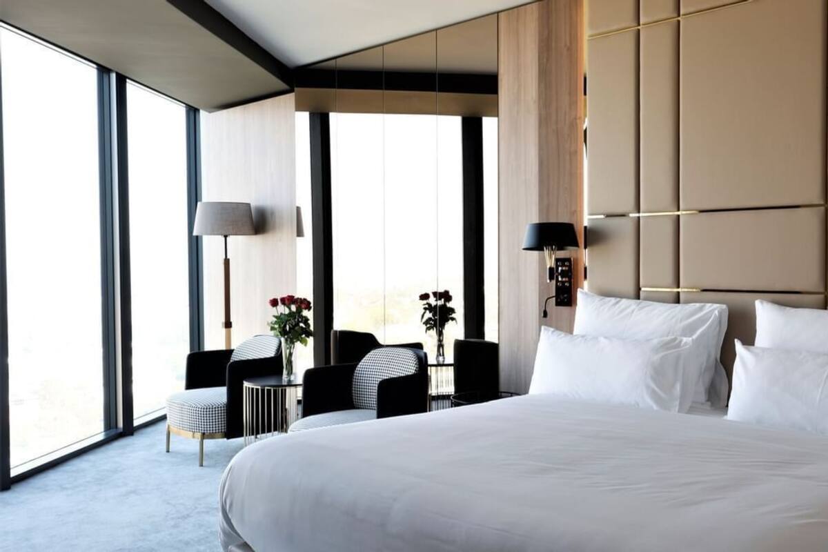 A large bed with white linens, a bedside table, a lamp, and a chair in a modern hotel room with large windows.