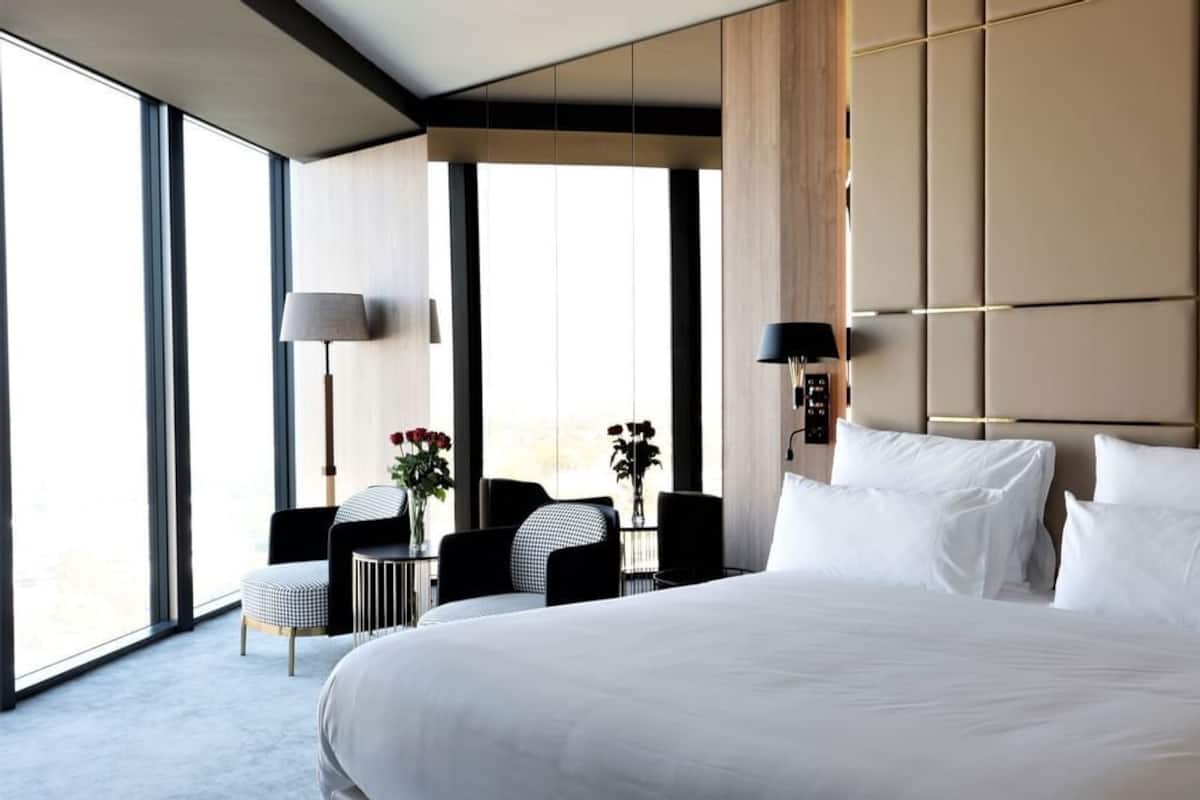 Una cama grande con sábanas blancas, una mesita de noche, una lámpara y una silla en una habitación de hotel moderna con ventanas amplias.