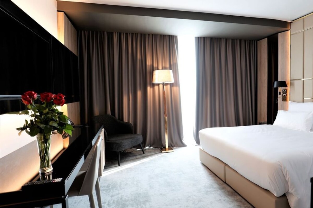 A hotel room with a bed, a desk with a lamp, a chair, and a vase of flowers.
