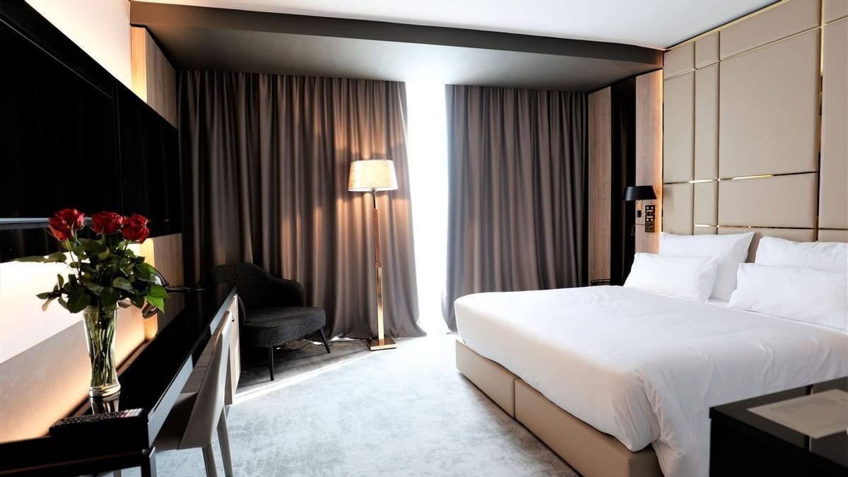 Una habitación de hotel con una cama, un escritorio con una lámpara, una silla y un florero con flores.