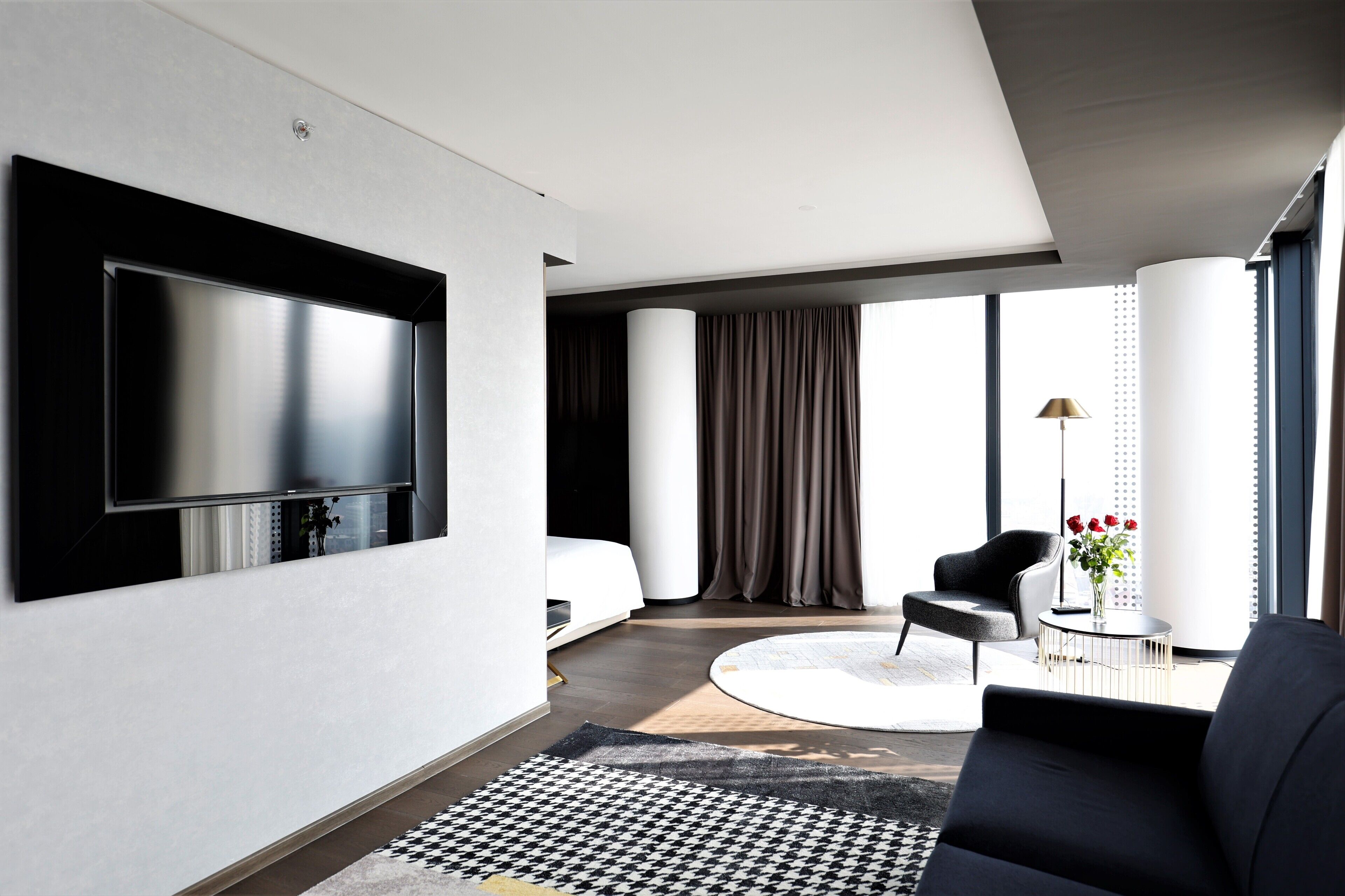 A modern hotel room with a large flat-screen TV mounted on the wall, a bed, a round coffee table with a vase of flowers, a black armchair, and a large window with curtains.
