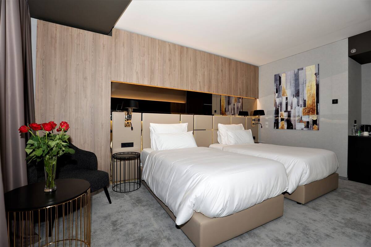 A modern hotel room with two beds, a small seating area, a vase with red flowers, and a large abstract painting on the wall.