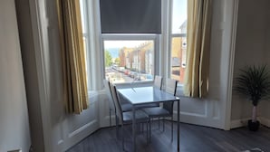 A corner room with a dining table and chairs, a potted plant, and a view of the street outside.