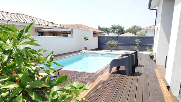 Um deck de madeira com piscina, uma pequena área de jardim e um edifício branco com telhado telhado.