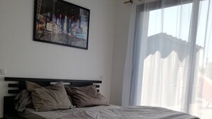 Quarto com cama, quadro na parede e janela com cortinas leves.