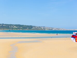 A beach with a clear sky, a sandy shore, and a distant view of a town with buildings. - The Sandbox (Hayle)