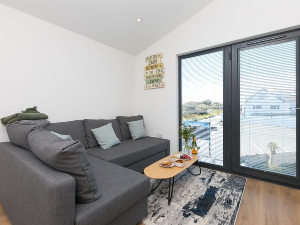 A modern living room with a grey sectional sofa, a small wooden table with snacks, a large window with blinds, and a motivational quote on the wall. - The Sandbox (Hayle)