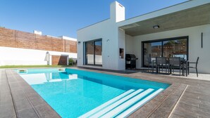 Un área de piscina moderna al aire libre con una terraza de madera, una parrilla y una zona de comedor con mobiliario exterior.