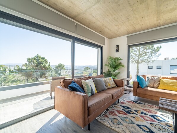 A modern living room with a brown leather sofa, a patterned rug, and large windows offering a view of the landscape.