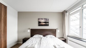 Ein ordentlich bezogenes Bett mit weißen Leinen und Handtüchern, ein hölzerner Kopfseitenbogen, ein Nachttisch mit Lampe sowie ein Gemälde mit einem Sonnenuntergang über Wasser an der Wand.