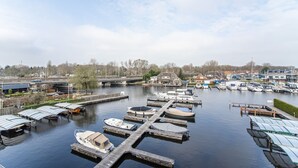 Ein Yachthafen mit an Stegen vertäuten Booten, eine Brücke im Hintergrund und eine Reihe Häuser entlang des Ufers.