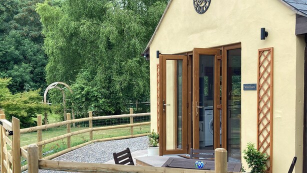 Une clôture en bois entoure un espace patio aménagé avec une table et des chaises, une horloge est fixée au bâtiment, et une enseigne indique « Mini Haven ».