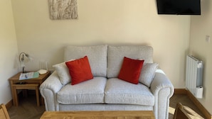 Un salon avec un canapé gris, deux coussins rouges, une table basse en bois, une petite table d’appoint avec une lampe, un tableau encadré au mur et un téléviseur fixé au mur.