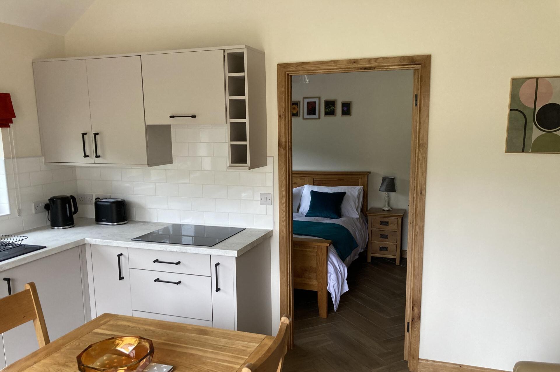Cozinha compacta com armários brancos, bancada e mesa de jantar com cadeiras. É visível, no quarto adjacente, uma cama com cabeceira de madeira e mesa de cabeceira com luminária.