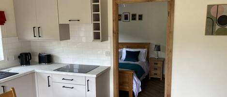 Cozinha compacta com armários brancos, bancada e mesa de jantar com cadeiras. É visível, no quarto adjacente, uma cama com cabeceira de madeira e mesa de cabeceira com luminária.