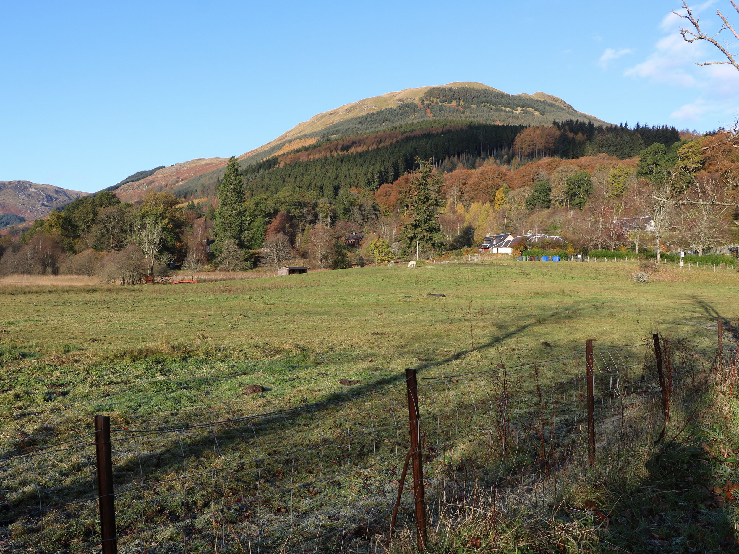 A rural landscape with a fenced field, a mountain in the background, and a few buildings.
