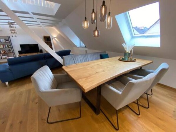 A modern loft-style room with a wooden dining table, two white chairs, a blue sofa, and a TV in the background.