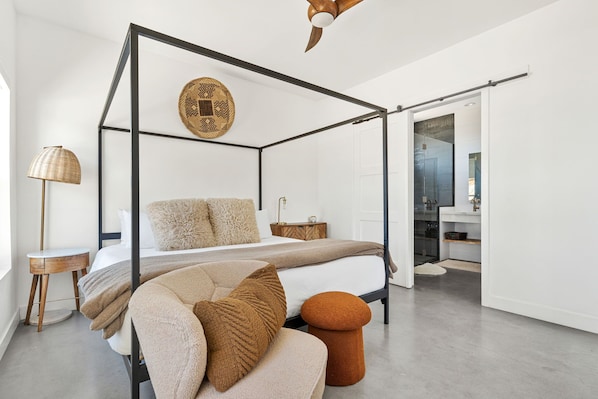 A modern bedroom with a four-poster bed, a round wall decoration, a bedside table, a lamp, and a view into the bathroom. - Contemporary Compound By Homestead Modern (Joshua Tree)
