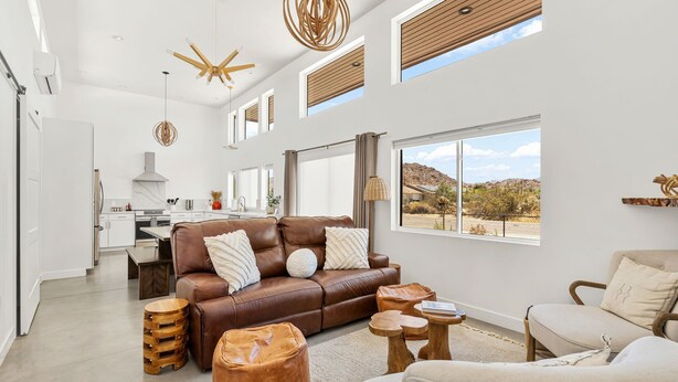 A spacious living area with a brown leather sofa, wooden ottomans, and a beige armchair. A kitchen is visible in the background, and large windows offer a view of the outdoors. - Contemporary Compound By Homestead Modern (Joshua Tree)