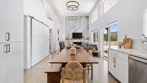A modern kitchen with a wooden dining table, white cabinetry, and a large window offering a view of the outdoors. - Contemporary Compound By Homestead Modern (Joshua Tree)