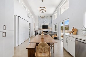 A modern kitchen with a wooden dining table, white cabinetry, and a large window offering a view of the outdoors. - Contemporary Compound By Homestead Modern (Joshua Tree)