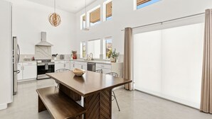 A modern kitchen with a large island, white cabinetry, and a large sliding glass door leading to a patio. - Contemporary Compound By Homestead Modern (Joshua Tree)