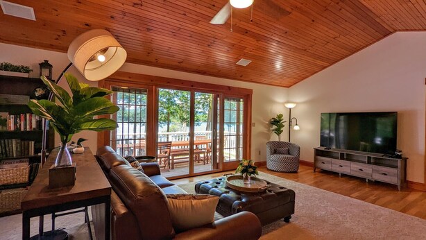 A spacious living room with a leather sofa, a coffee table, and a TV. There is a bookshelf and a dining area visible through large sliding glass doors. - Hideaway at Highland Lake (Andover)