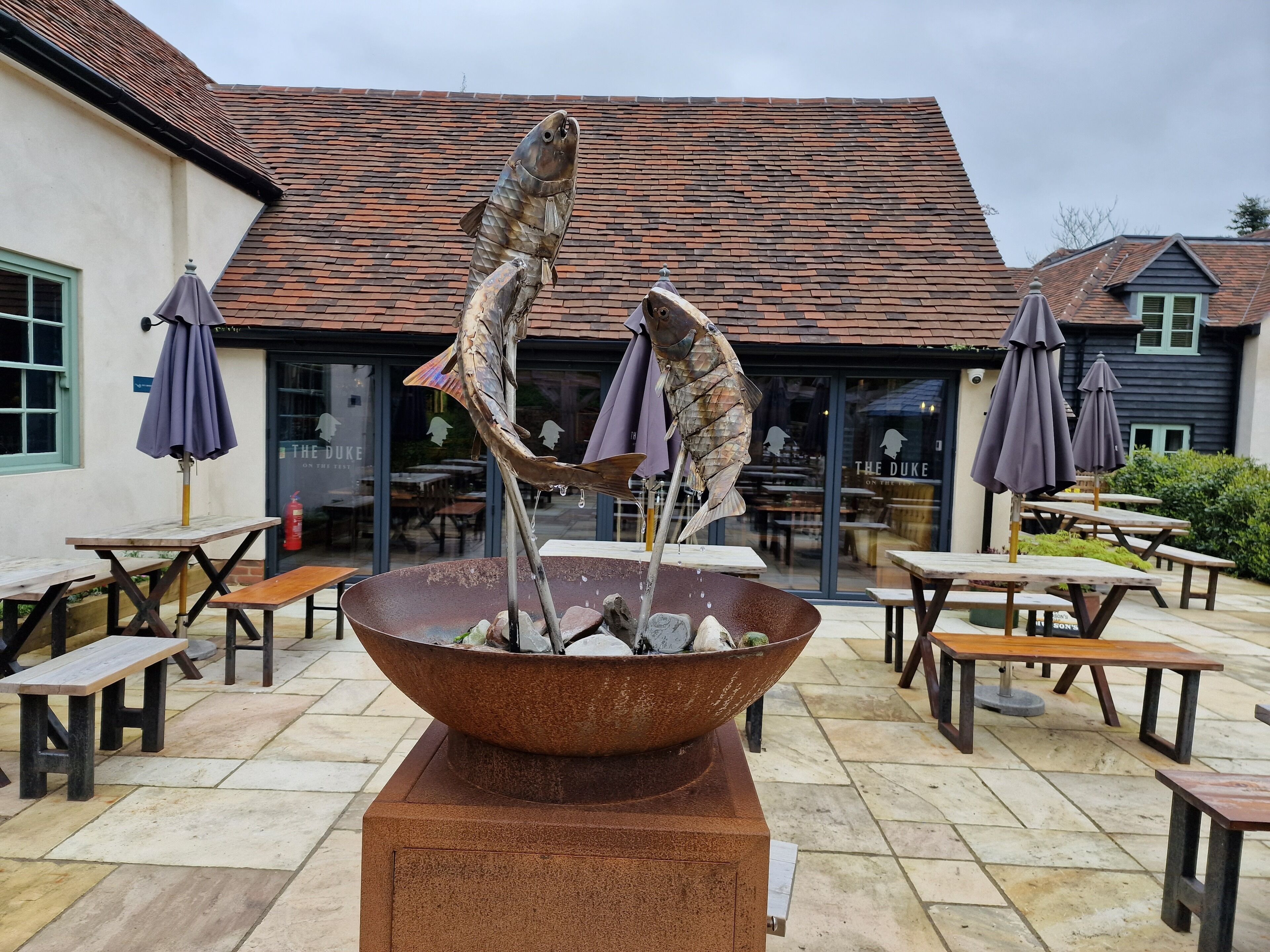 An outdoor dining area with a central fish sculpture, wooden tables, and umbrellas.