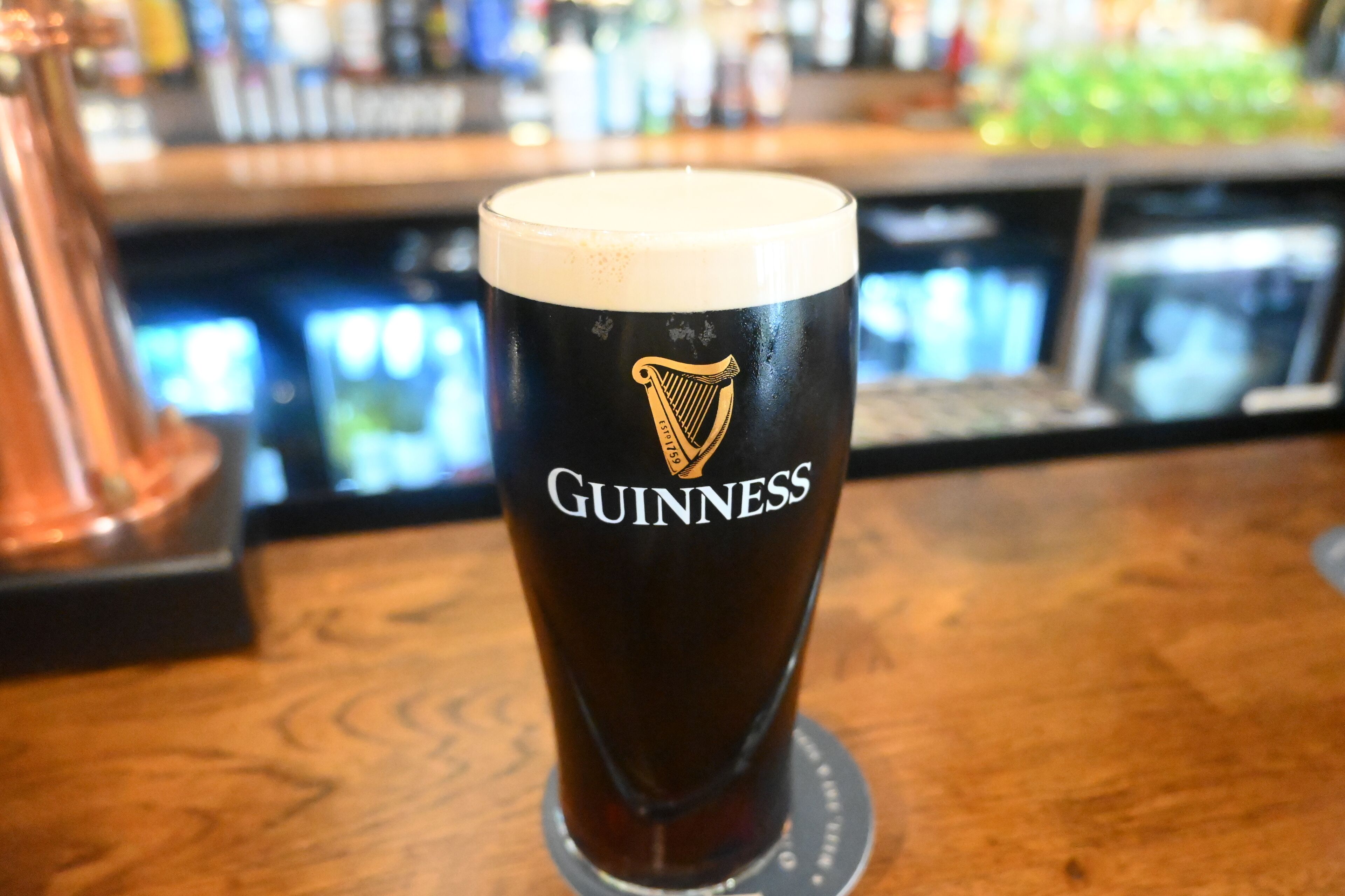 A Guinness glass on a wooden bar counter with a blurred background of bottles and bar equipment.