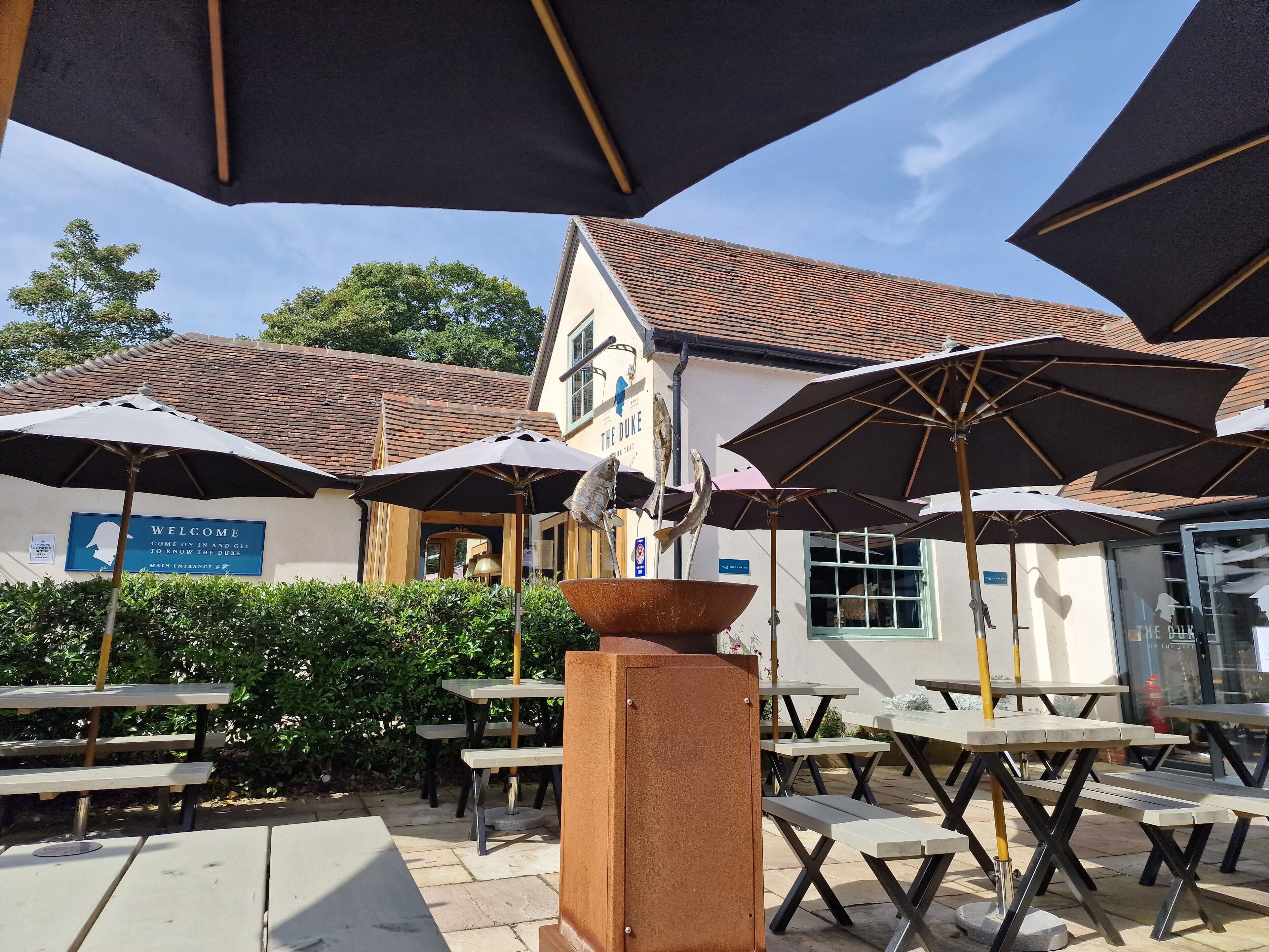 Un espace extérieur aménagé avec des tables et des parasols, une enseigne accueillant les visiteurs et un bâtiment doté d’un toit en tuiles.