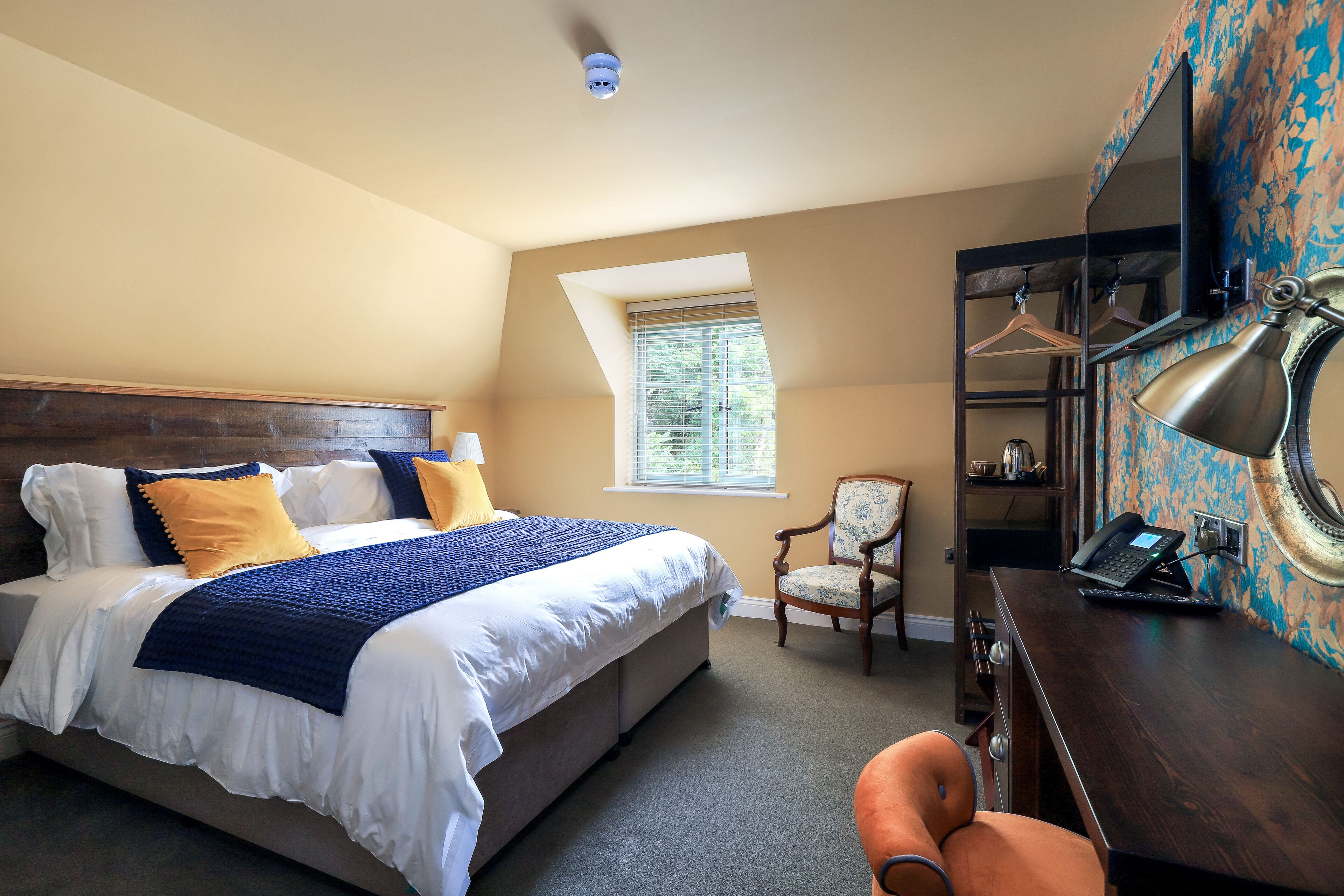 Une chambre avec un grand lit, un bureau avec un ordinateur portable, une chaise, un porte-manteau et une fenêtre avec des stores.
