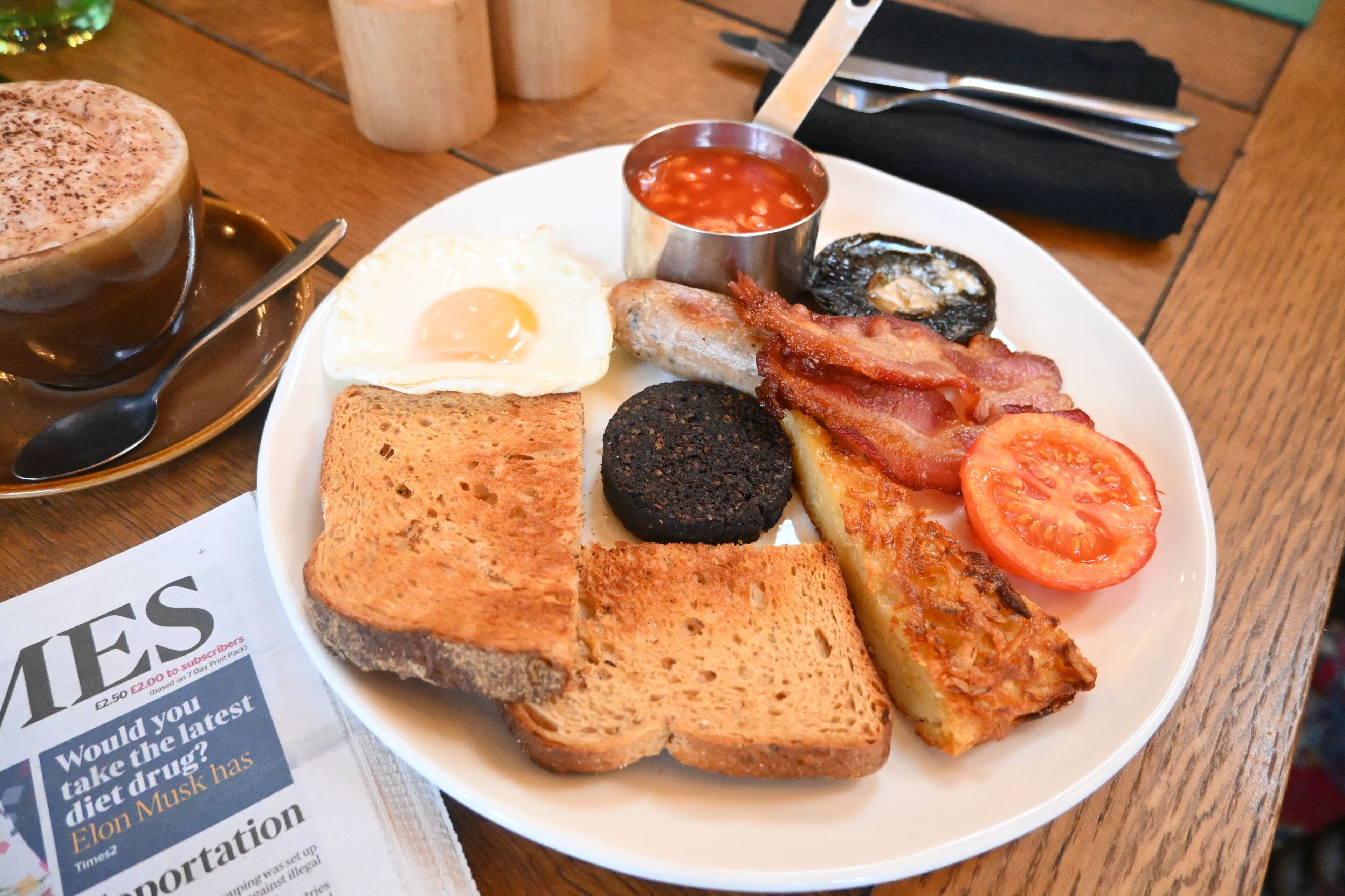 A breakfast plate with toast, eggs, bacon, black pudding, a slice of tomato, and a small container of ketchup.