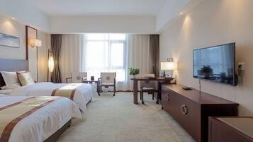 A hotel room with two beds, a desk, a chair, a TV, and a large window with curtains.