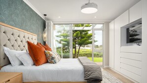 Un dormitorio con una cama grande, una mesita de noche de madera, un televisor montado en la pared y vistas a zonas verdes a través del ventanal.