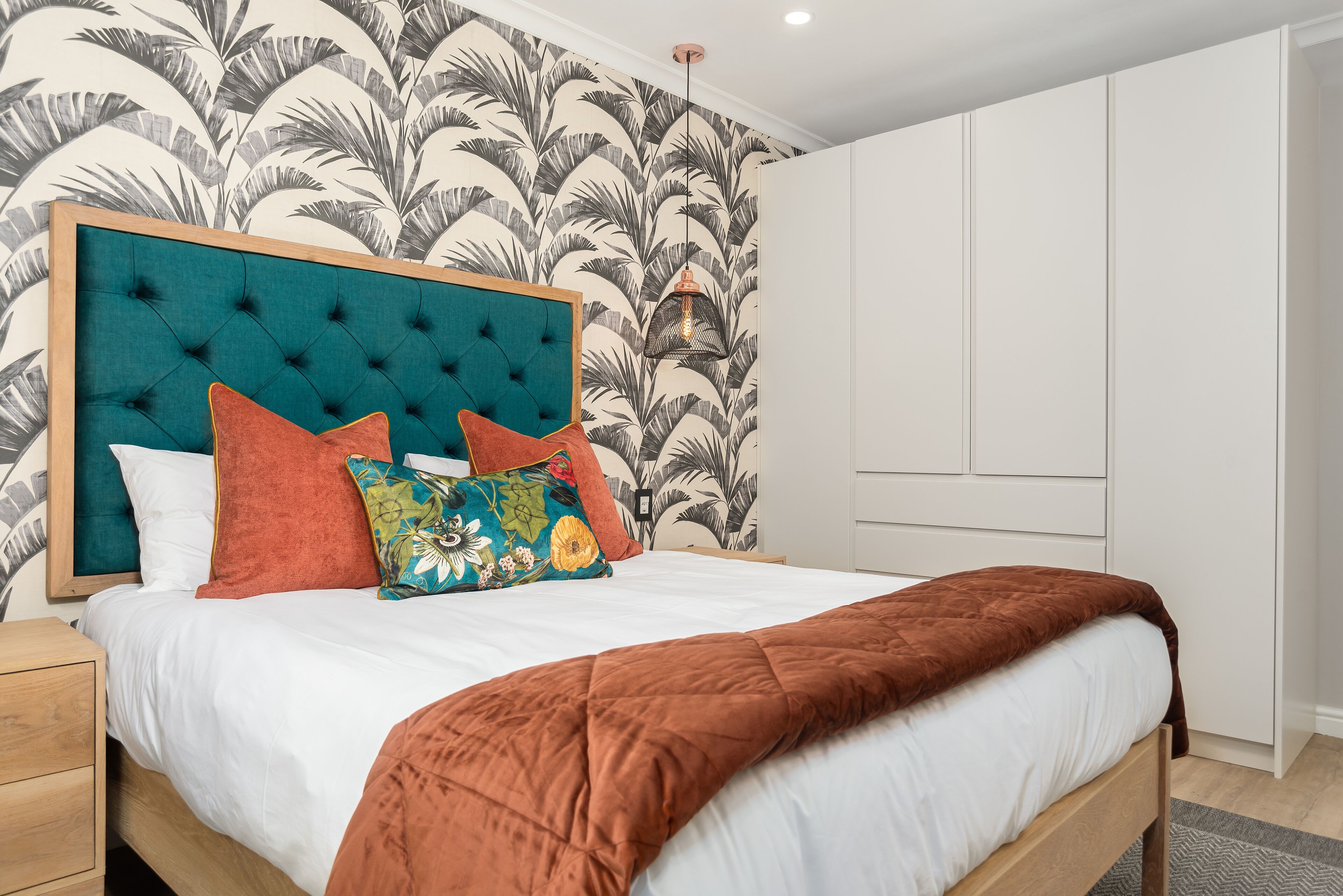 Een slaapkamer met een gestoffeerd hoofdeinde, een bed met wit beddengoed en een bruine plaid, kleurrijke kussens, en een muur met een tropisch bladmotief.