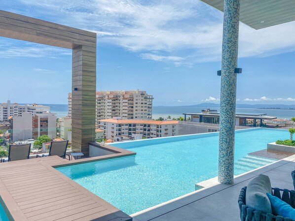 Ein Pool auf dem Dach mit klarem, blauem Wasser, umgeben von modernen Gebäuden und Blick auf das Meer.