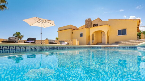 Une piscine avec un bord carrelé, un espace de détente avec des chaises et un parasol, et un bâtiment doté d’un toit et d’une cheminée recouverts de tuiles.