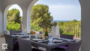 A dining area with wicker furniture, a table set for four, and a view of the sea and trees. - Villa CORASOL by Abahana Villas (Teulada)