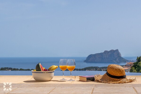 A table with a bowl of fruit, two glasses of orange juice, a straw hat, and a book, overlooking a coastal view with a mountain in the distance.