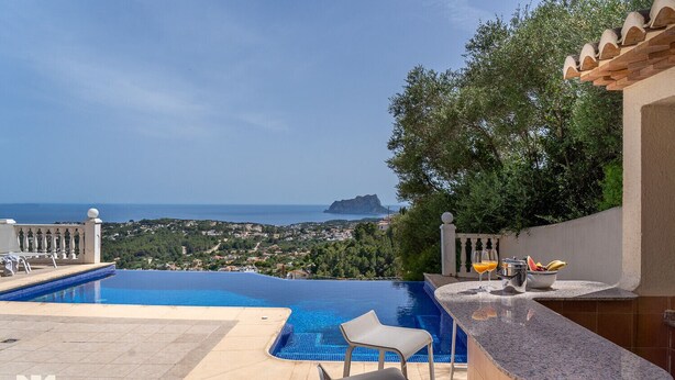 An infinity pool with a view of the sea and a distant island, a bar area with seating, and a lush green backdrop. - Villa TREVERIS by Abahana Villas (Teulada)