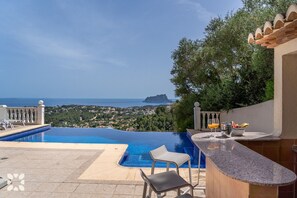 An infinity pool with a view of the sea and a distant island, a bar area with seating, and a lush green backdrop.