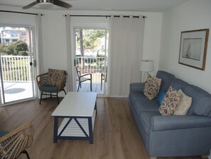 A living room with a sofa, a table, and chairs. There is a balcony with a railing and a view of the outside.
