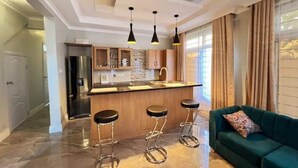 Eine moderne Küche mit einer zentralen Kochinsel, Barhocker, Pendelleuchten und einem grünen Sofa mit einem gemusterten Kissen.