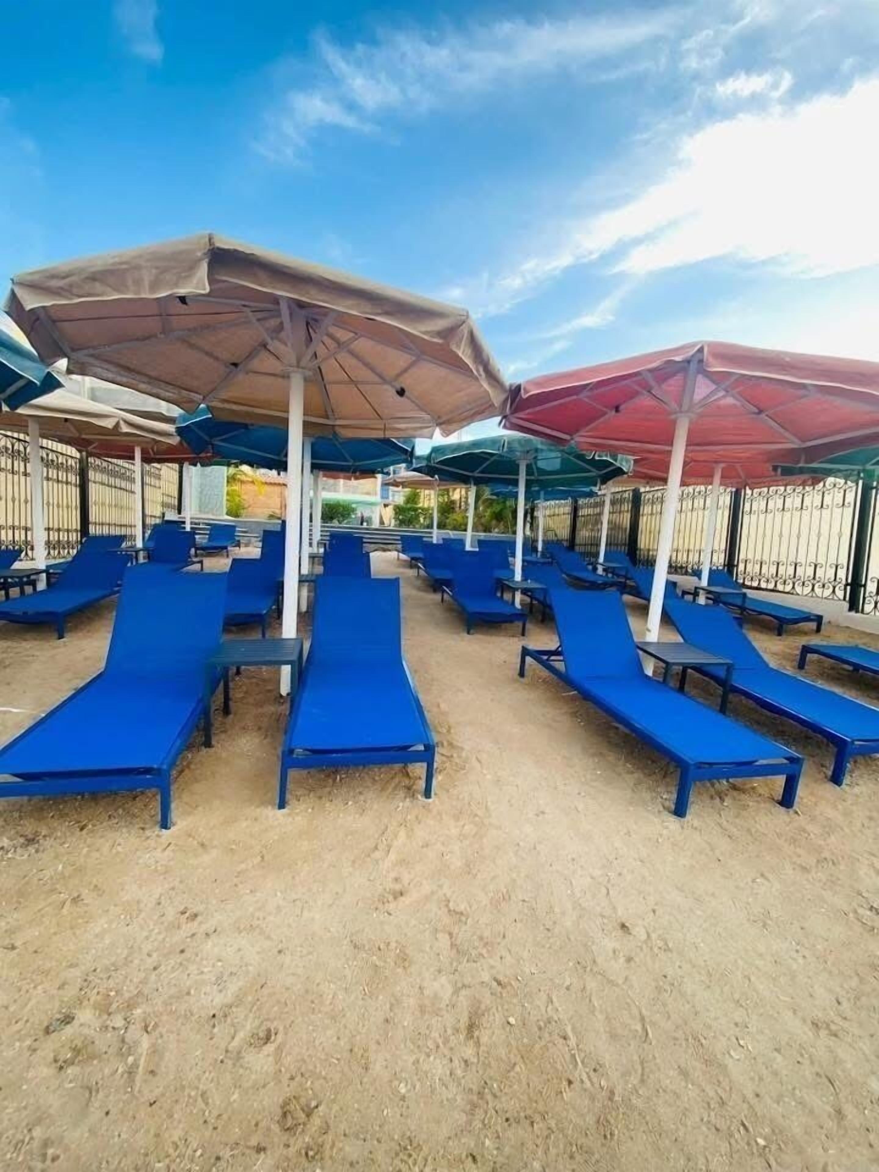 Ein Strandbereich mit blauen Liegestühlen und Sonnenschirmen, sandigem Untergrund und blauem Himmel.