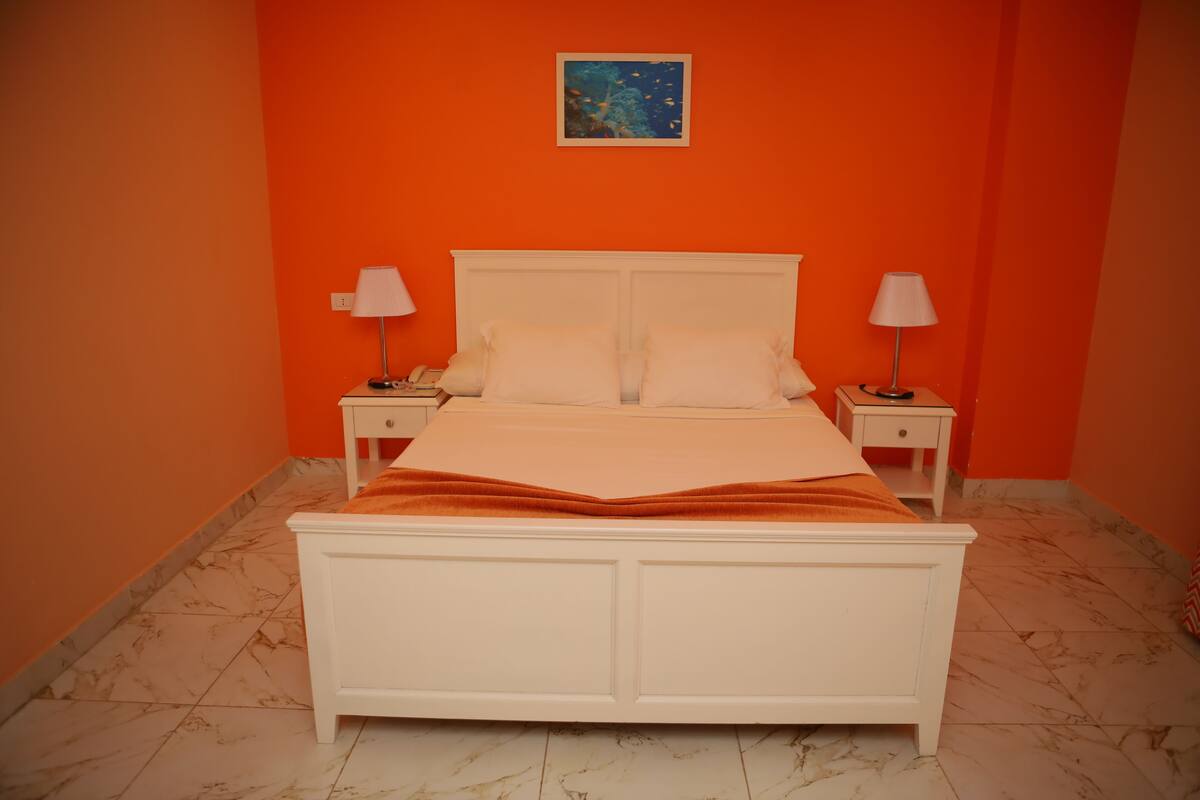 Ein Schlafzimmer mit einem weißen Bett, orangefarbenen Wänden und einem Nachttisch mit einer Lampe auf jeder Seite.