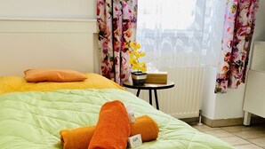 Hotelkamer met een groene plaid, oranje kussens, bloemen gordijnen en een tafeltje met een bloevas.
