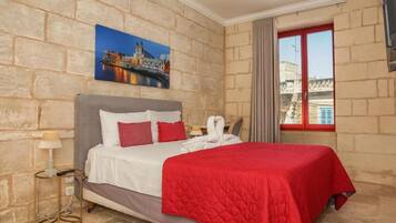 A hotel room with a stone wall, a bed with a red bedspread, a painting of a cityscape, a window with curtains, and a view of buildings outside.
