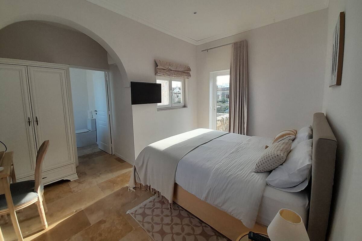 A bedroom with a bed, a TV mounted on the wall, a wardrobe, and a window with curtains.