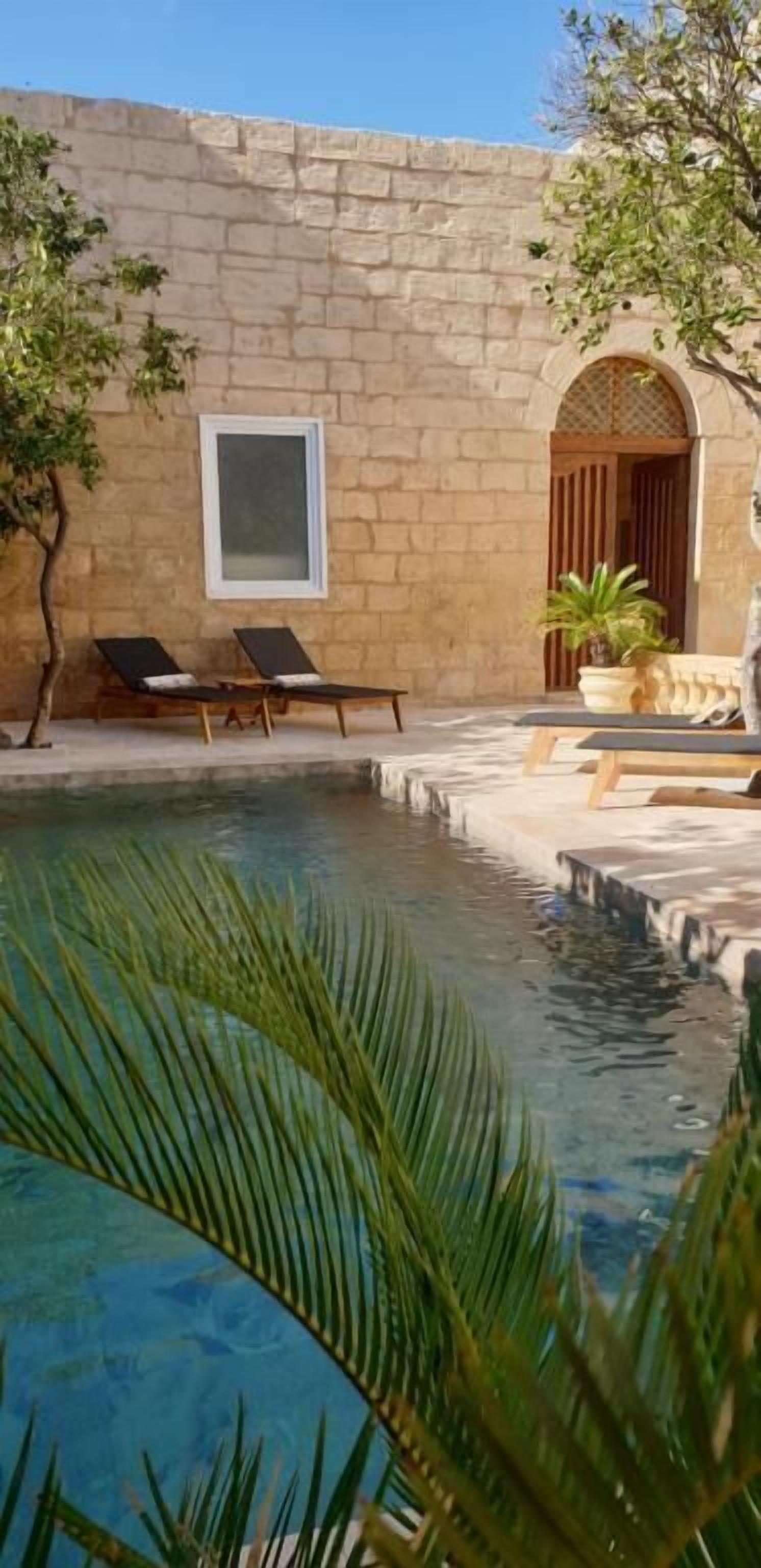 A stone building with a pool, two lounge chairs, and a potted plant.