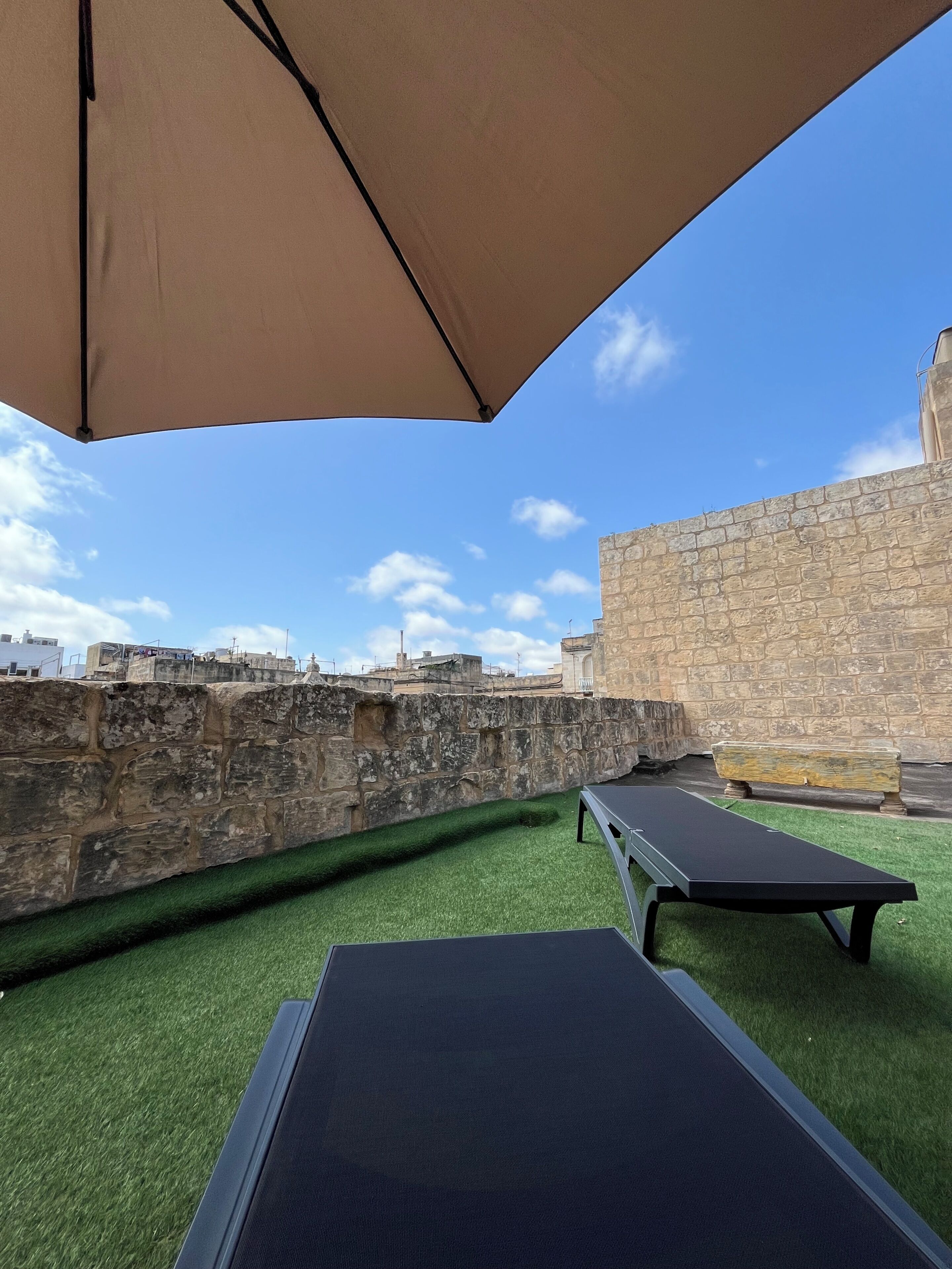 An outdoor area with a stone wall, green artificial turf, and two lounge chairs under a large umbrella.